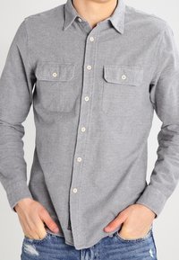 Mężczyzna ubrany w szarą koszulę na guziki z dwoma kieszeniami na piersi oraz niebieskie, postrzępione dżinsy, ręce wsunięte w przednie kieszenie.