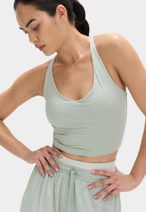 Młoda kobieta w jasnooliwkowym bezrękawniku typu crop top i pasujących spodniach ze sznurkiem, ręce spoczywają na biodrach, patrzy w dół w lewo.