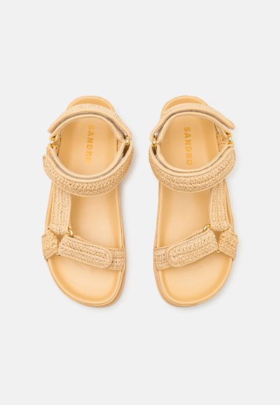 Beige sandaler med vevde stropper, med to justerbare spenner og en glatt såle. Merket "SANDRO" trykt på innsiden.