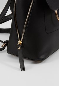 Gros plan sur le coin d'un sac à main en cuir noir avec une fermeture éclair dorée et une languette de tirage, montrant la sangle et les détails de couture arrondis.