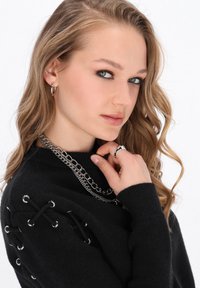 Schwarzer Pullover mit Schnürdetails und silbernen Ösen. Das Model trägt geschichtete silberne Ketten und einen Ring. Hellbraunes Haar, das in Wellen gestylt ist.