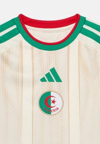 Maillot de sport beige et crème à rayures avec le logo Adidas vert et l'emblème national algérien comprenant un croissant rouge et une étoile.