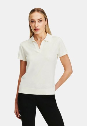Donna con capelli biondi lisci indossa una polo bianca a maniche corte e pantaloni neri, in piedi con una mano sui fianchi.