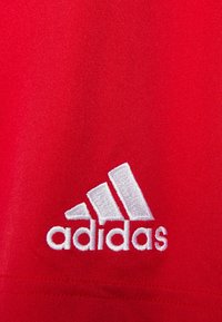 Camiseta deportiva roja con un logo de Adidas bordado en blanco en el lado izquierdo. La tela tiene una textura suave y es liviana.