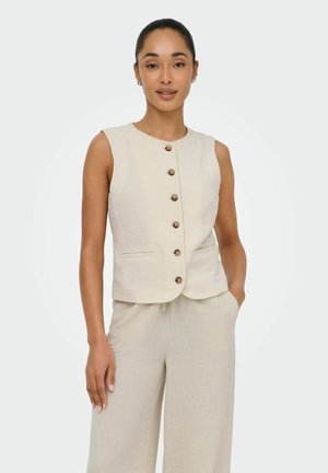 Femme portant un gilet beige sans manches boutonné et un pantalon à jambes larges assorti, une main dans la poche, debout devant un fond uni.