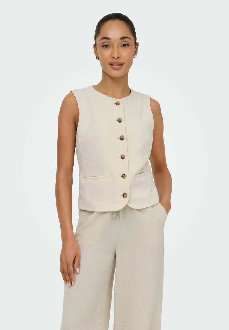 Femme portant un gilet beige sans manches boutonné et un pantalon à jambes larges assorti, une main dans la poche, debout devant un fond uni.
