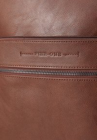 Sac en cuir marron avec une surface texturée, comportant une poche zippée et le logo "PIER ONE" en relief au-dessus de la fermeture éclair. Design simple et fonctionnel.