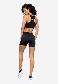 Top crop atletico nero con texture a coste e design racerback, abbinato a pantaloni corti neri a vita alta. Il modello indossa scarpe da ginnastica nere.