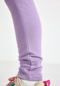 Μωβ κολάν κατασκευασμένο από μαλακό, ελαστικό ύφασμα, με εφαρμογή που αγκαλιάζει το σώμα και ορατές ραφές, συνδυασμένο με πολύχρωμα αθλητικά παπούτσια.