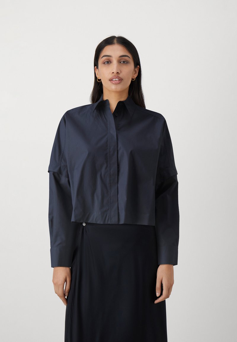 Camicetta navy cropped realizzata in tessuto morbido con chiusura a bottoni, colletto ampio e maniche lunghe, abbinata a una gonna scura dal design minimalista.