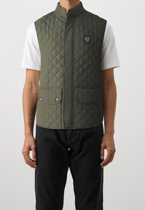 Homme portant un gilet sans manches matelassé vert olive avec des poches pression, par-dessus une chemise blanche à manches courtes et un pantalon noir, debout devant un fond uni.