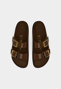 Καφέ σανδάλια Birkenstock με δύο λαμπερές ρυθμιζόμενες λουρίδες και χρυσά αγκράφες, εμφανίζονται από πάνω σε λευκό φόντο.