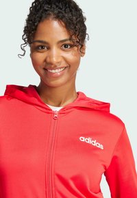 Červená mikina na zips s bielym logom "adidas" na ľavej hrudi. Obsahuje kapucňu so šnúrkou a hladkú, elastickú textúru materiálu.