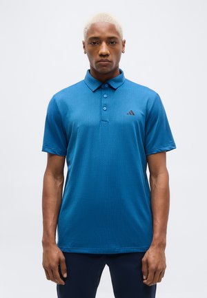 Rövid szőke hajú férfi kék texturált rövid ujjú galléros pólót viselve, amelyen kis logó található a mellrészen, fehér háttér előtt áll.