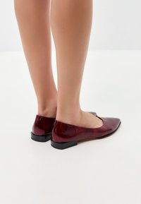 Ballerines en cuir verni rouge avec un design épuré, un bout pointu et un petit talon noir, mettant en valeur une texture brillante et des détails subtils.