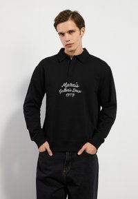 Svart polo-lignende hettegenser med krage, med hvit brodert tekst "Marais Gallerie Deux 1973" på brystet, og ribbestrikkede mansjetter.