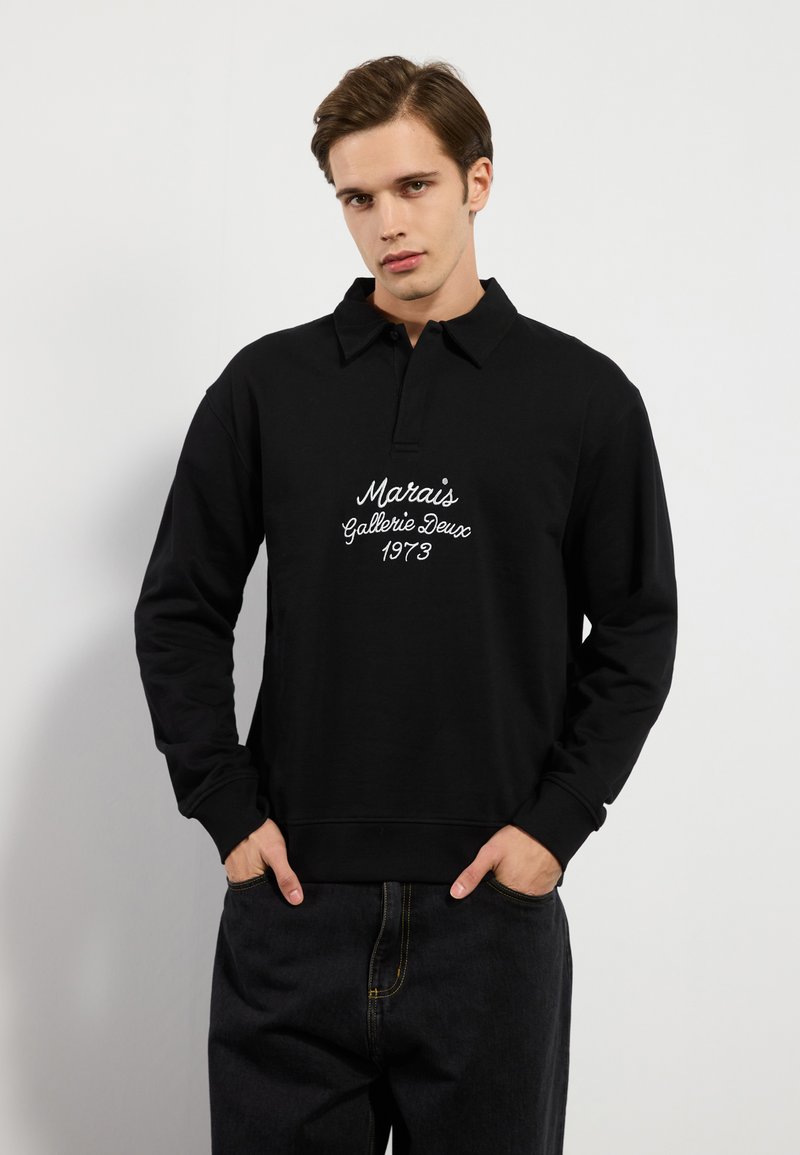 Svart polo-lignende hettegenser med krage, med hvit brodert tekst "Marais Gallerie Deux 1973" på brystet, og ribbestrikkede mansjetter.