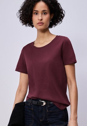 Jeune femme aux cheveux bouclés portant un t-shirt bordeaux à manches courtes, rentré dans un jean foncé avec une ceinture noire.