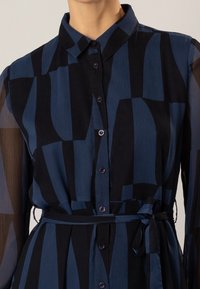 Blue and black patterned shirt dress with a button-up front, collar, long sheer sleeves, and a tied waist belt. Textured fabric.