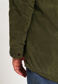 Olivgrön jacka med mjuk textur, med knäppta ärmslut och en avslappnad passform. Kjolen är något förlängd bak.