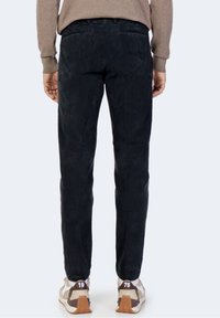 Dunkle Cordhose mit geripptem Muster und schmaler Passform, ausgestattet mit zwei Gesäßtaschen, kombiniert mit beigen Sneakers und einem beigefarbenen Pullover.