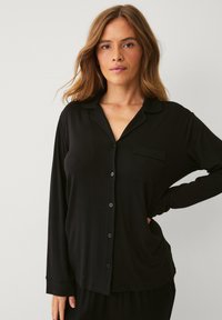 Sort langærmet pyjamasoverdel med knaplukning fortil, reverskrave og en enkelt brystlomme, lavet af blødt stof med en glat tekstur.