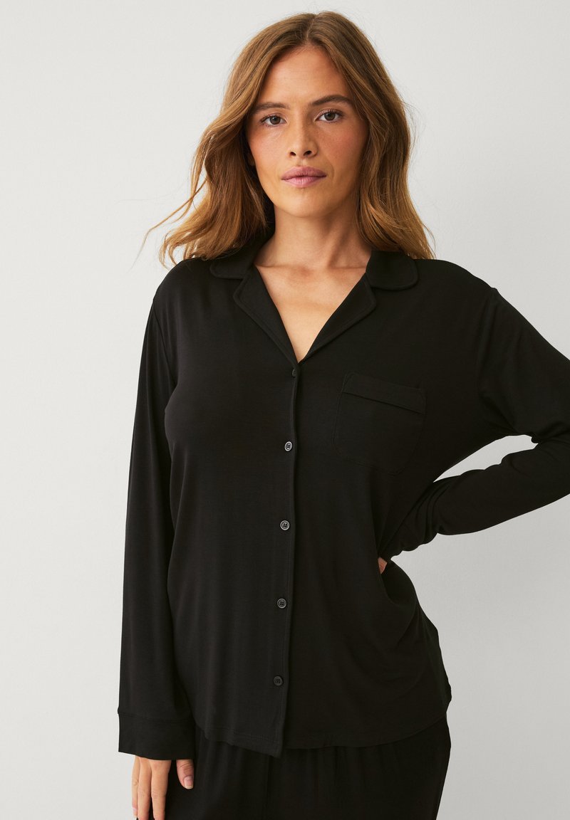 Sort langærmet pyjamasoverdel med knaplukning fortil, reverskrave og en enkelt brystlomme, lavet af blødt stof med en glat tekstur.