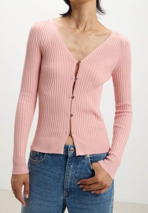 Person iført en tætsiddende ribbet lyserød cardigan med knapper kombineret med blå jeans, hånd hvilende nær lommen, neutralt baggrund.