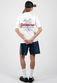 Άτομο με λευκό γραφικό T-shirt με κόκκινο και μπλε κείμενο και ιπτάμενο άλογο, ναυτικό ριγωτό σορτς, λευκές κάλτσες, μαύρα παπούτσια και μαύρο καπέλο, όρθιο με τα χέρια πίσω από την πλάτη.