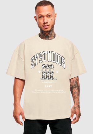 Beige übergroßes T-Shirt mit kräftigem schwarzen Text "2Y STUDIOS WORLDWIDE 1992" und einem grafischen Design, das drei Figuren darstellt. Baumwollmaterial, entspannte Passform.
