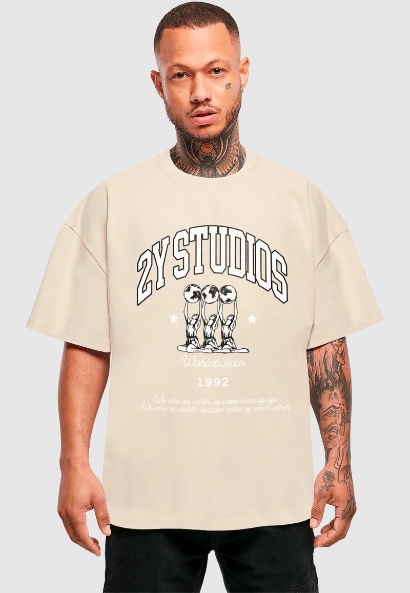 Camiseta oversize beige con texto negro en negrita "2Y STUDIOS WORLDWIDE 1992" y diseño gráfico que representa tres figuras. Material de algodón, corte relajado.