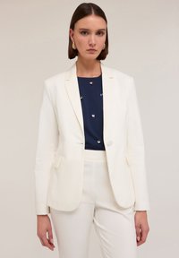 Blazer blanc ajusté avec revers crantés et fermeture �à un bouton, associé à une chemise bleu marine ornée d'accents métalliques et à un pantalon clair.