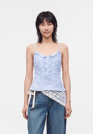 Junge Frau mit kurzem schwarzen Haar trägt ein hellblau gestreiftes ärmelloses Oberteil und lockere blaue Jeans mit einem weißen Strickschal, der in der Taille gebunden ist.