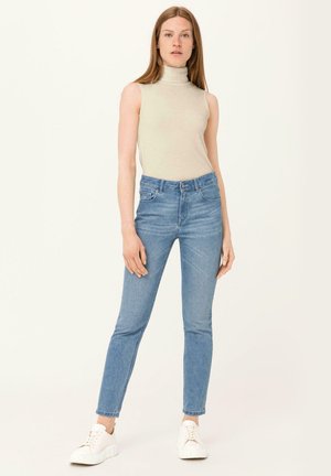 Lichtbeige mouwloze coltrui gecombineerd met hoge taille blauwe denim jeans en witte sneakers, met een nauwsluitend ontwerp en taps toelopende benen.