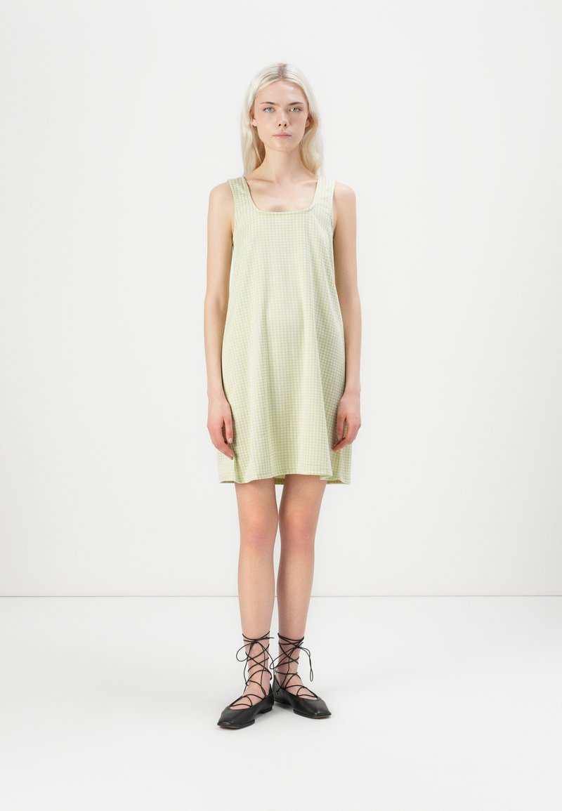 Vestido sem mangas com padrão xadrez verde e decote quadrado, feito de tecido leve; modelo a usar sapatos pretos com atacadores.
