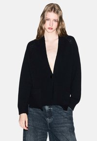 Jonge vrouw met lang haar die een zwarte gebreide cardigan met een diepe V-hals en losse, vervaagde blauwe jeans draagt tegen een witte achtergrond.