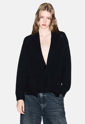 Junge Frau mit langen Haaren, die eine schwarze Strickjacke mit tiefem V-Ausschnitt und lässigen, hellblauen Jeans trägt, vor einem weißen Hintergrund.