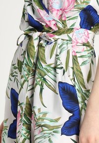 Vestido envolvente floral en tejido ligero. Presenta flores en tonos rosa y azul, hojas verdes y un cinturón ajustable. Fondo claro con textura suave.