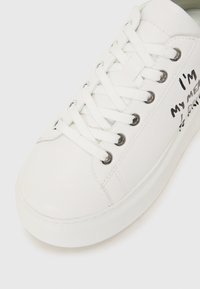 White sneaker with smooth leather material, round toe, flat rubber sole, silver eyelets, and black printed text on the side.