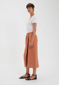 Jupe midi en terre cuite en tissu léger, avec une fente frontale et un design plissé, associée à des sandales noires à lanières et un haut blanc.