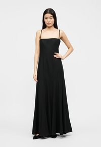 Sieviete stāv, turot roku uz gurna, tērpusies garā melnā kleitā ar plāniem siksniņām un melnās smailās kurpēs uz vienkrāsaina fona.