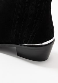 Czarne zamszowe botki za kostkę z niskim, drewnianym obcasem i srebrnym wykończeniem nad obcasem na białym tle.