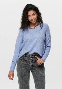Pull en tricot côtelé bleu clair avec une coupe décontractée et des manches longues, associé à un jean noir à taille haute en délavé acide et un collier en chaîne argentée.