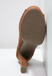 Zapato de tacón alto de ante marrón con diseño perforado, punta afilada y suela de goma texturizada con ranuras horizontales.