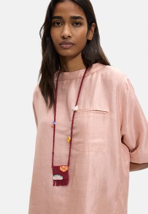 Kobieta o długich ciemnych włosach nosi luźną, jasnoróżową bluzkę i długi koralikowy naszyjnik z małymi, kolorowymi owocowymi i chwostowymi detalami.