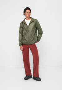 Olivgrön zip-up-jacket med en lätt, blank yta, i kombination med löst sittande röda byxor. Outfiten har en enkel vit t-shirt under.