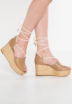 Jambes portant des sandales compensées en cuir beige avec semelles plateformes tressées et liens blancs à nouer autour de la cheville, debout sur un sol blanc.