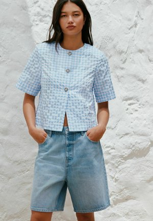 Frau, die ein hellblaues, kurzärmeliges Hemd mit vichy-Karos und lockere Jeansshorts trägt, steht vor einer weißen, strukturierten Wand.