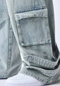 Pantalon cargo en denim bleu clair avec de grandes poches latérales et un ourlet effiloché, associé à une paire de baskets blanches. La texture est légèrement usée.
