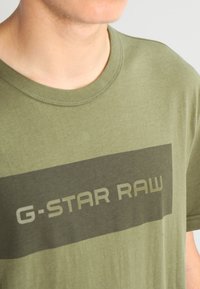 Camiseta de algodón verde oliva con cuello redondo, que presenta un gráfico rectangular oscuro con el texto "G-STAR RAW" en gris claro.